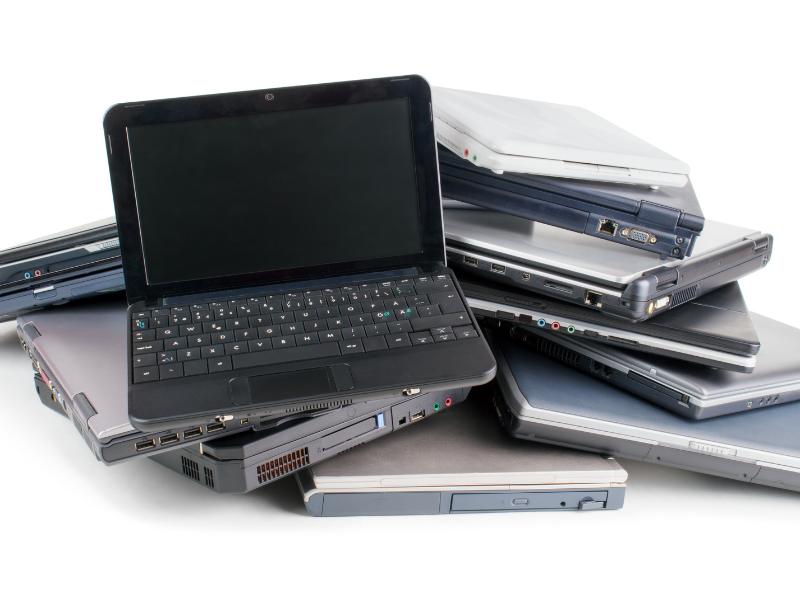 Pile of old laptops stacked on a white background, with one open black laptop in front and several closed silver and gray notebooks behind.